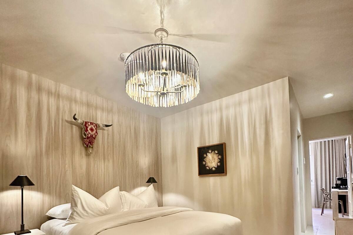 Une chambre à coucher avec un grand lit, une table de chevet avec une lampe, un lustre, une décoration murale représentant une tête de taureau et un tableau encadré.
