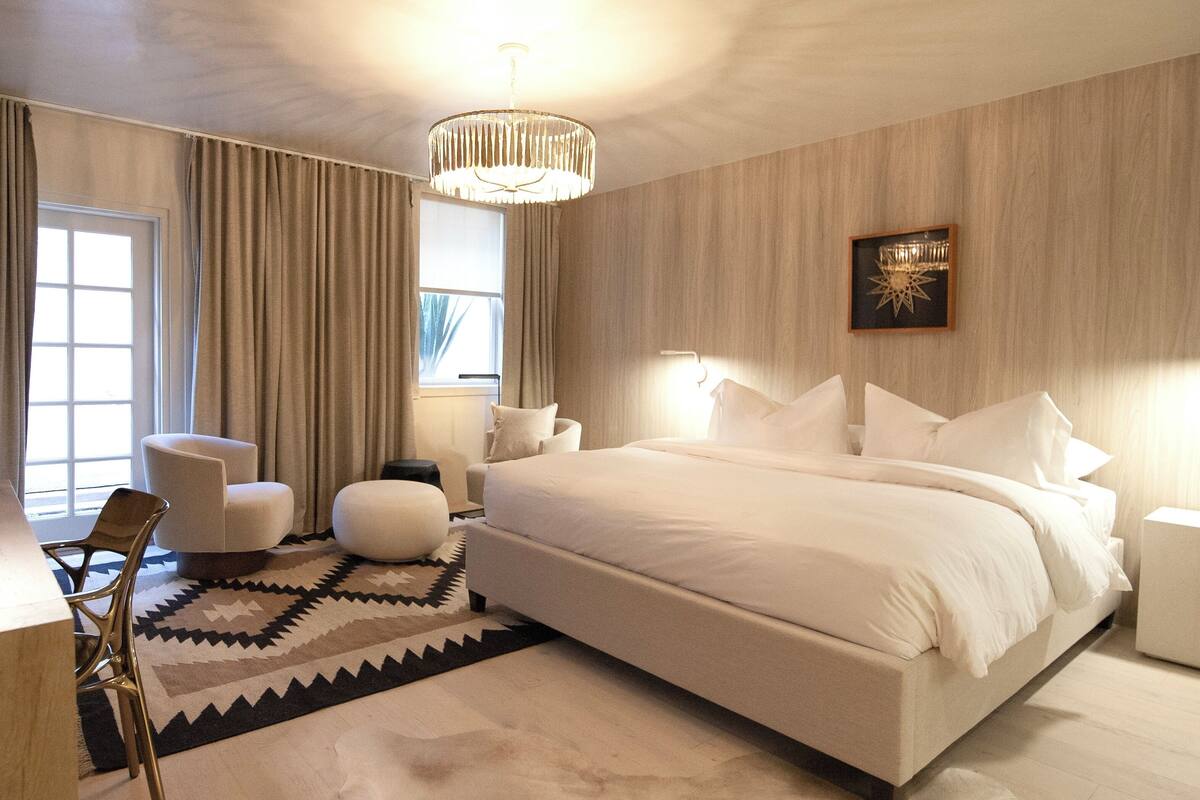 Une chambre moderne avec un grand lit, un coin salon comprenant un fauteuil et un pouf, un lustre et une fenêtre avec des rideaux.