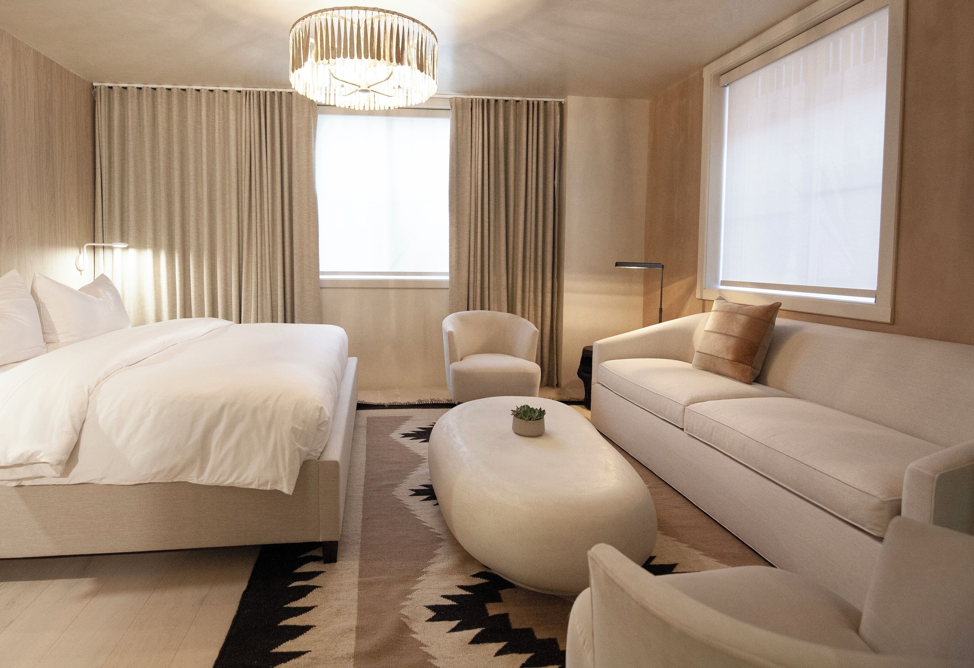 Habitación de hotel moderna con cama, sofá, mesita de centro y un amplio ventanal con cortinas.