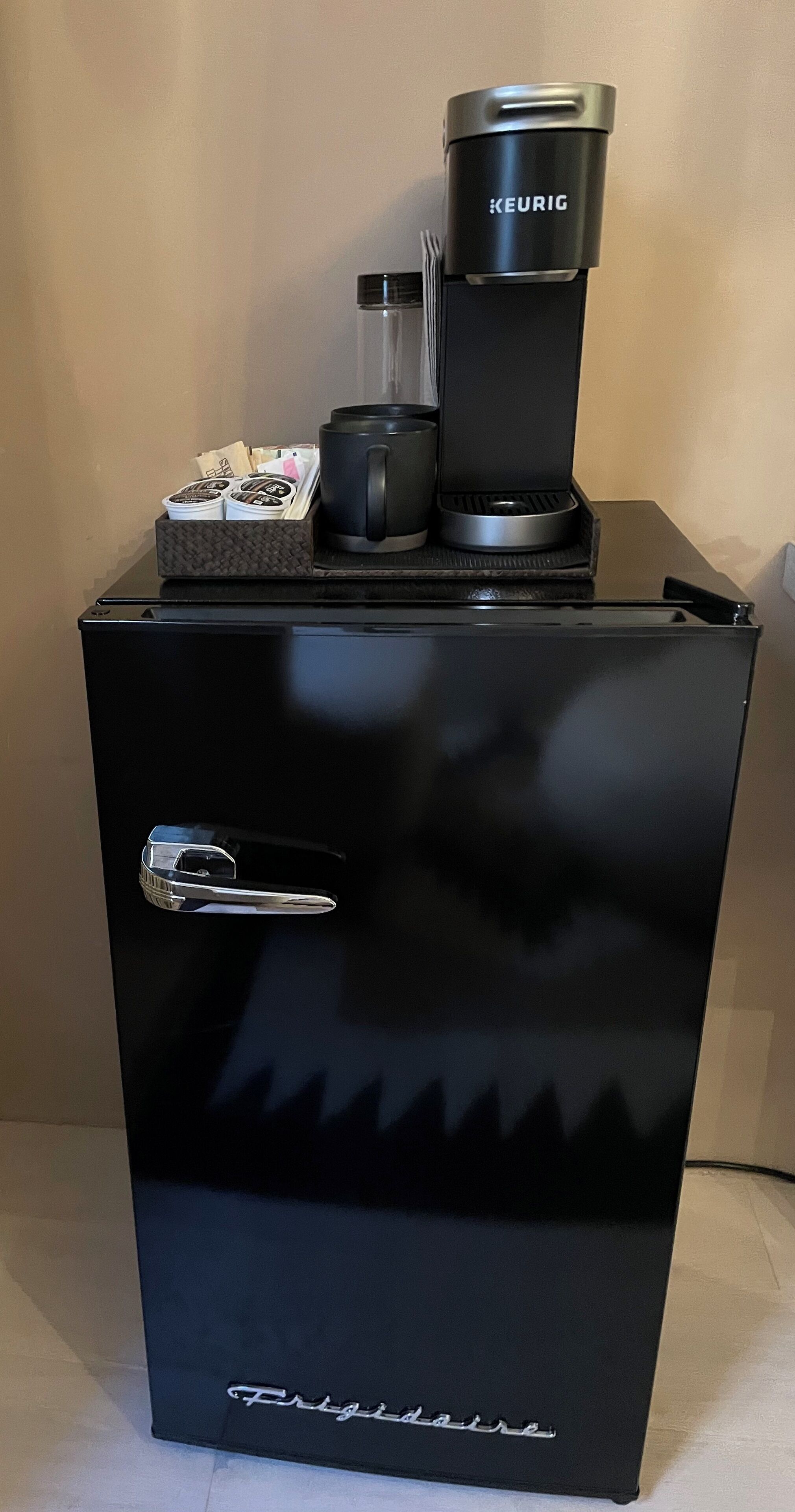 Ett svart kylskåp med en KEURIG-kaffebryggare ovanpå, ett litet torkställ och en hållare för kaffekapslar.