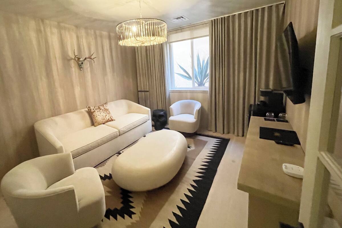 Un salon moderne avec un canapé blanc, deux fauteuils blancs, une table basse, une tête de cerf accrochée au mur, un lustre, un téléviseur et une fenêtre avec des rideaux.