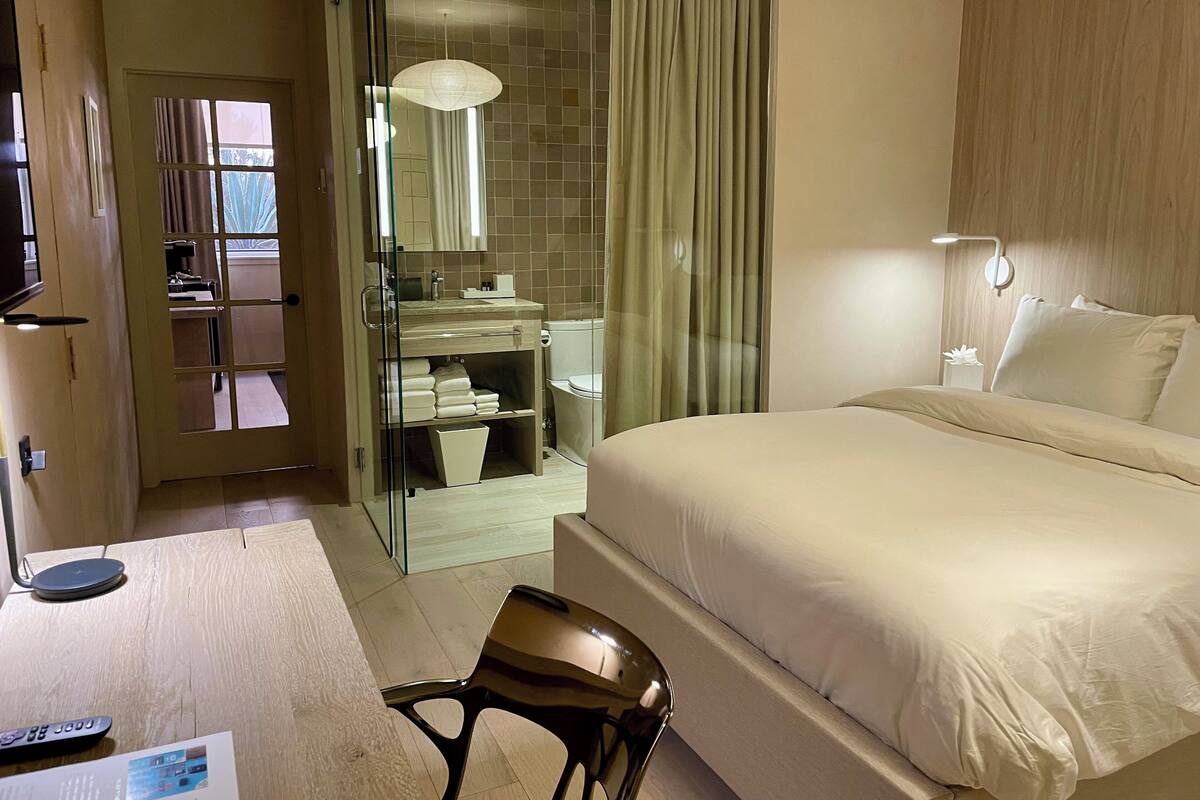 Une chambre d’hôtel avec un grand lit, un bureau avec une chaise, une salle de bain fermée par des parois de verre et une table de chevet avec une lampe.