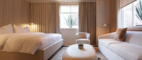 A modern hotel room with a large bed, a sofa, a round ottoman, and a chandelier.