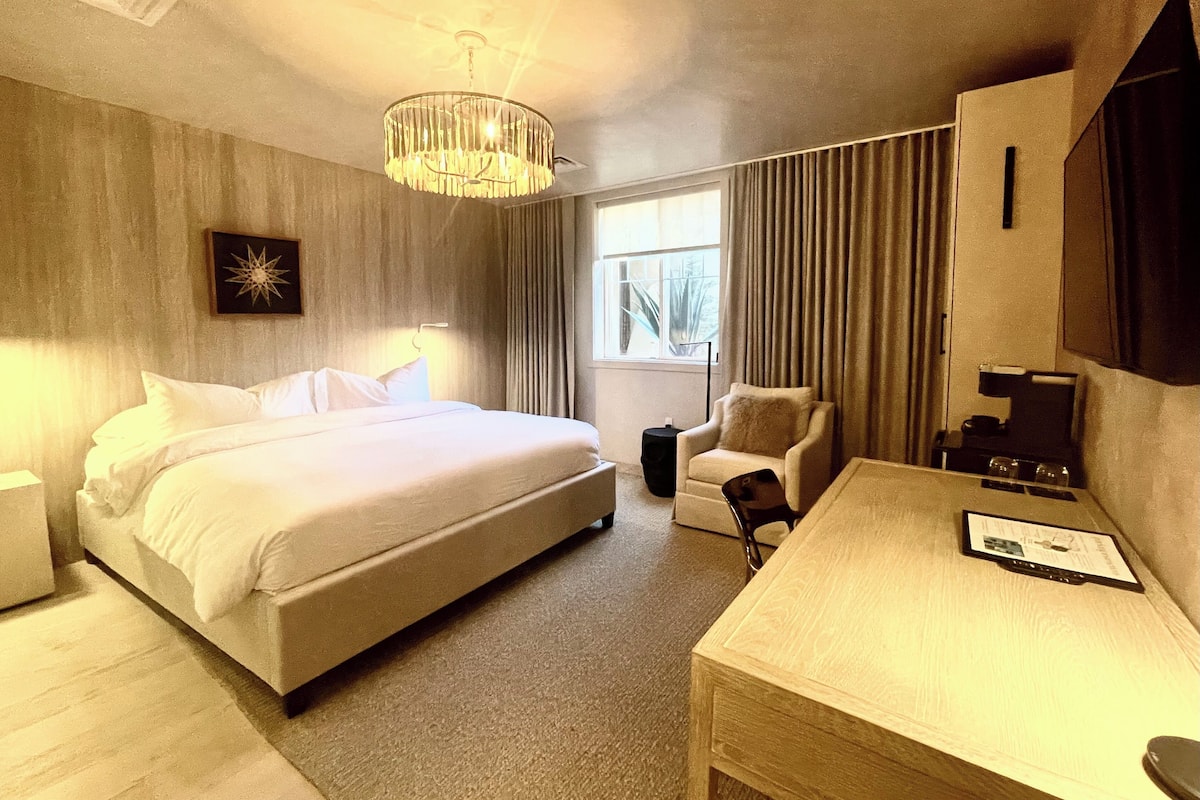 Une chambre d’hôtel avec un grand lit, un bureau, une chaise, une lampe de bureau, une petite table, une horloge, un cadre photo, une fenêtre avec des rideaux et un lustre.