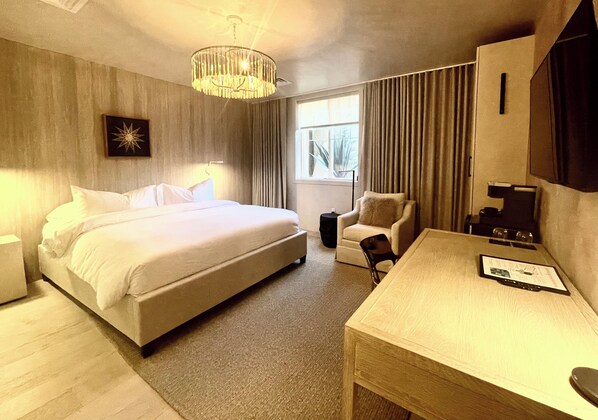 Habitación de hotel con una cama grande, un escritorio, una silla, una lámpara de escritorio, una mesita, un reloj, un marco de fotos, una ventana con cortinas y un candelabro.