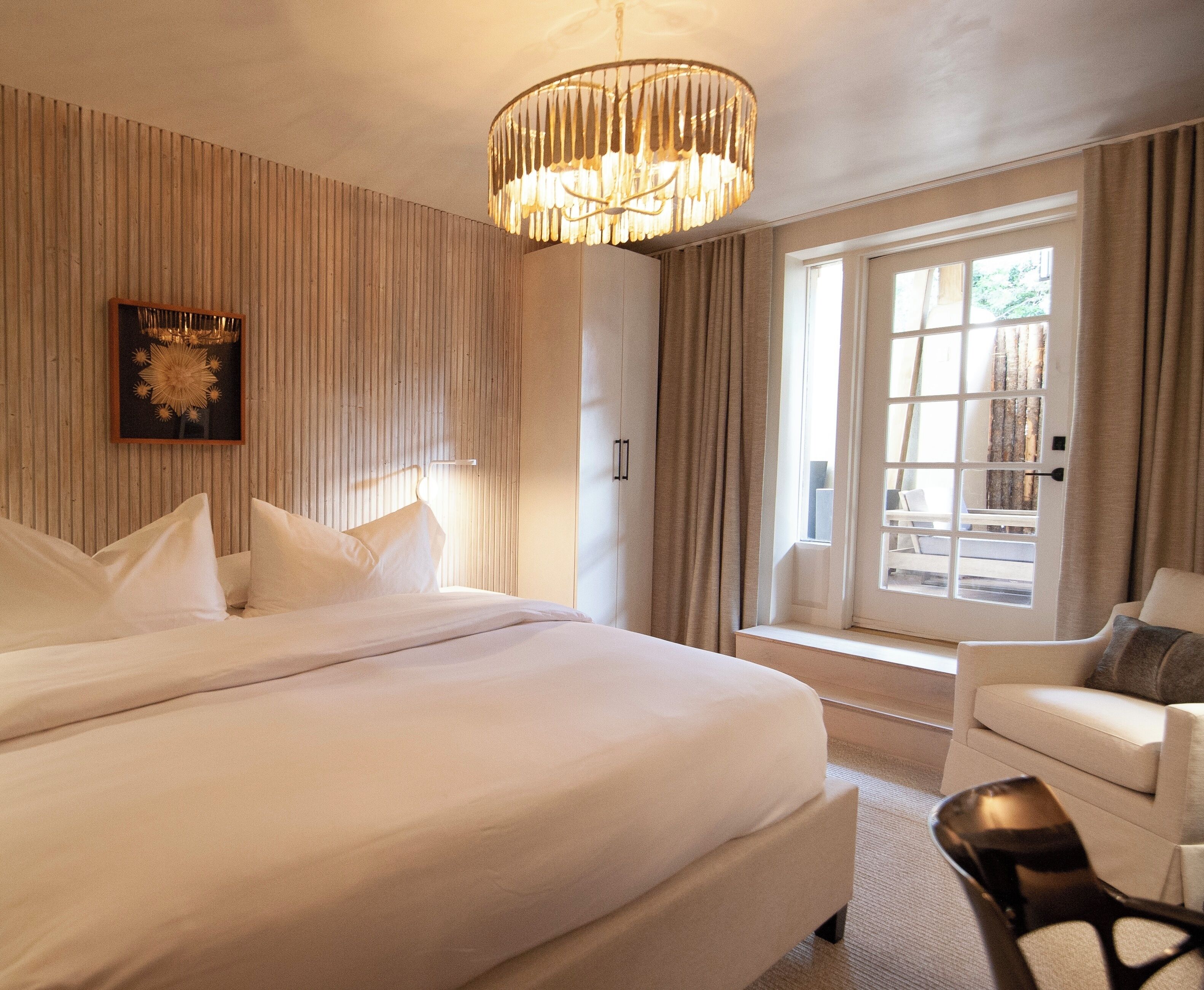 A hotel room with a large bed, a chair, a window with curtains, and a chandelier.