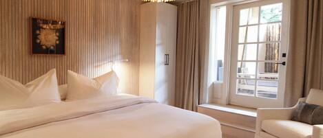 A hotel room with a large bed, a chair, a window with curtains, and a chandelier.