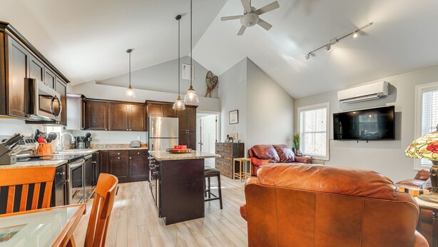 A spacious kitchen with dark wooden cabinets, a central island, and modern appliances. There is a large brown leather sofa in the living area, a dining table with chairs, and a ceiling fan.
