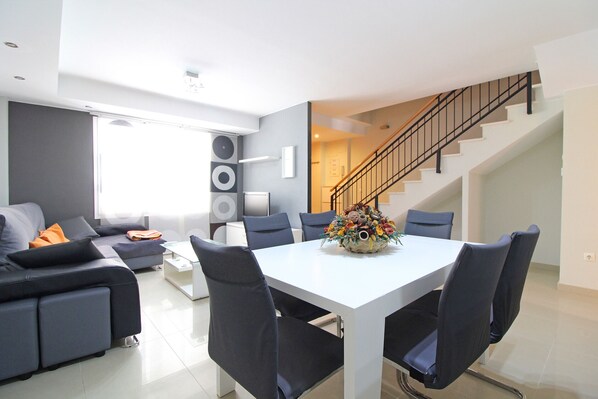 A modern living room with a white dining table, dark chairs, a sofa, and a staircase.