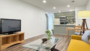 A modern living room with a flat-screen TV on a wooden stand, a glass coffee table with a decorative bowl, a yellow sofa, and a dining area with a wooden table and chairs. - Spacious Entire Home in Great Location ! (Chamblee)