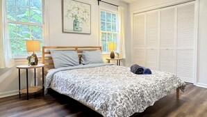 A bedroom with a wooden bed, two bedside tables with lamps, a large window with curtains, a closet with louvered doors, and a floral painting on the wall. - Spacious Entire Home in Great Location ! (Chamblee)