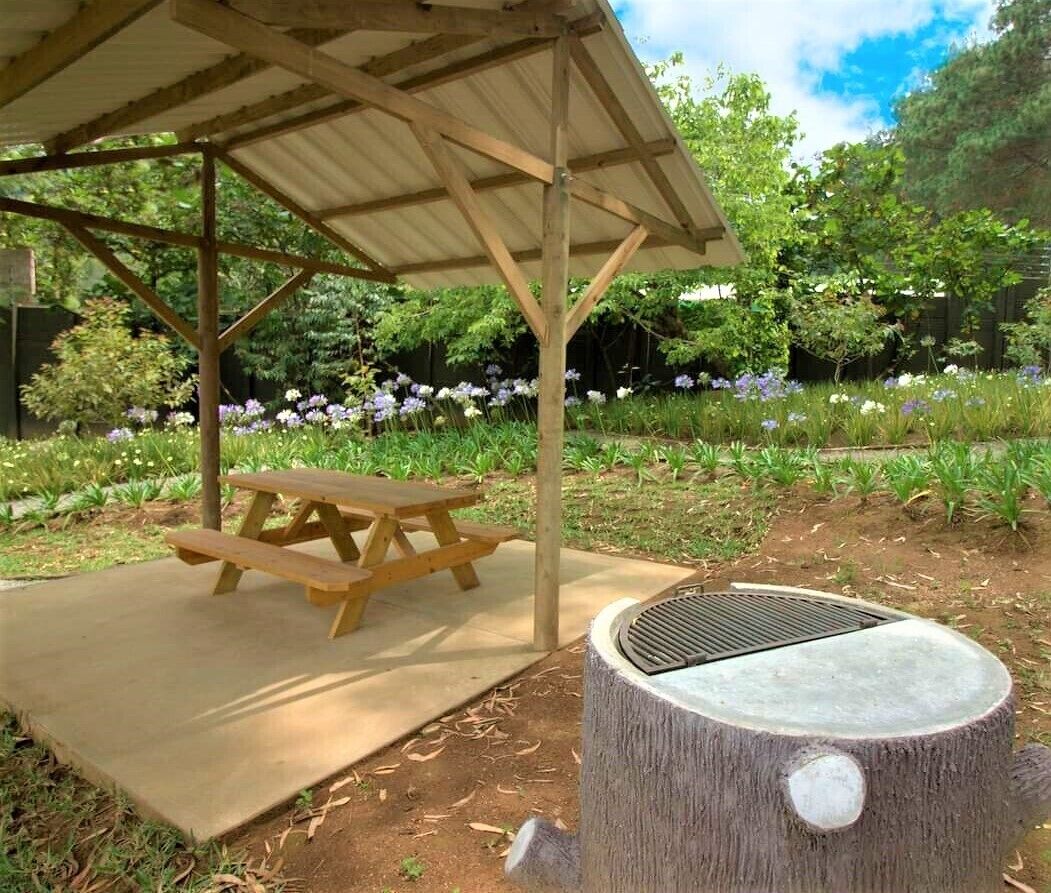A picnic area with a wooden table and bench, a covered shelter, and a tree stump with a grill.