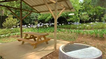 A picnic area with a wooden table and bench, a covered shelter, and a tree stump with a grill.