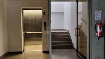An interior corridor with a glass door leading to an elevator, a fire extinguisher mounted on the wall, and a staircase with a fire exit sign.