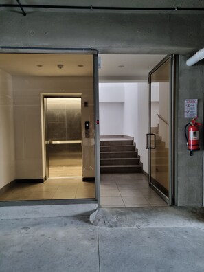 An interior corridor with a glass door leading to an elevator, a fire extinguisher mounted on the wall, and a staircase with a fire exit sign.