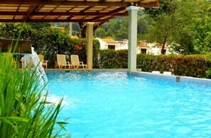 A swimming pool with clear blue water, surrounded by greenery and a covered area with chairs.
