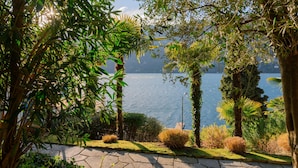 A landscaped garden with a stone pathway leading to a body of water, surrounded by various trees and shrubs. - La Casa - Spacious apartment in historic villa with private lake access (Carate Urio)