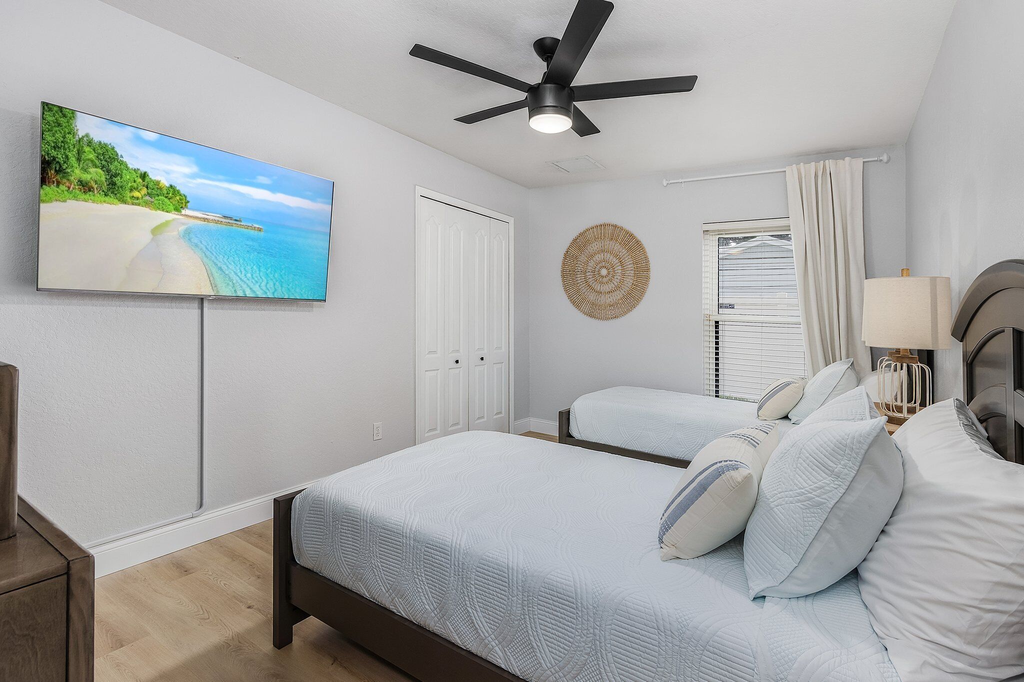 A bedroom with two beds, a ceiling fan, a TV on the wall, and a window with curtains.