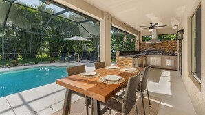 Une terrasse extérieure avec une piscine, une table à manger, des chaises et un coin cuisine doté d’un mur de protection en pierre.