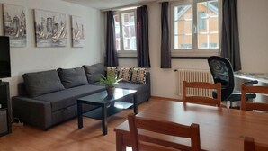 A modern living room with a sofa, coffee table, dining table, and chairs. There are three windows with curtains, a radiator, and artwork on the walls. - Charming 2-bedroom apartment with WiFi in St. Gallen (St. Gallen)