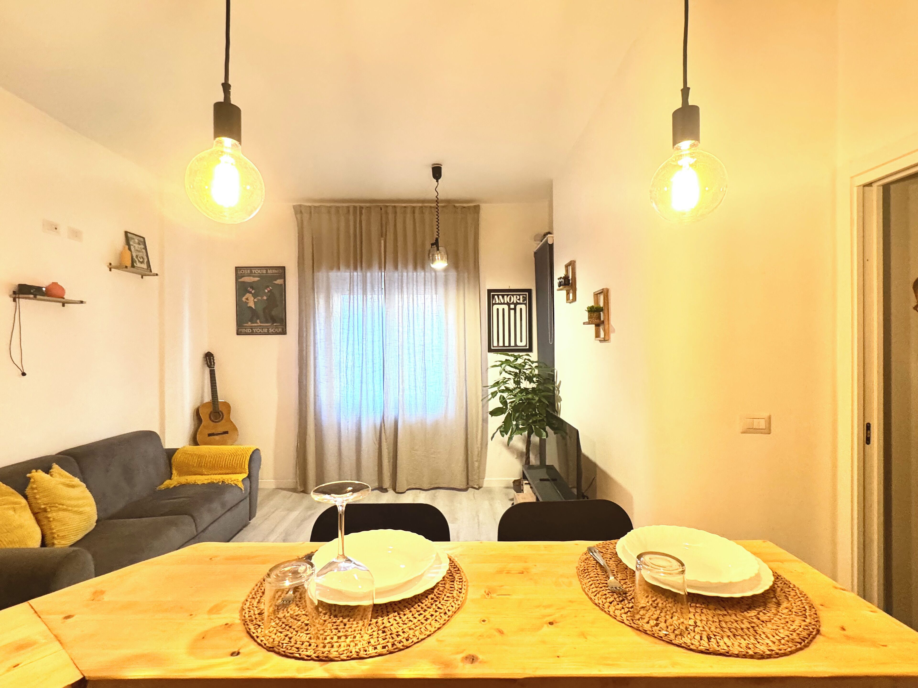 Un salon chaleureux avec un canapé, une table à manger dressée avec des assiettes et un verre à vin, une guitare et une grande fenêtre avec des rideaux.