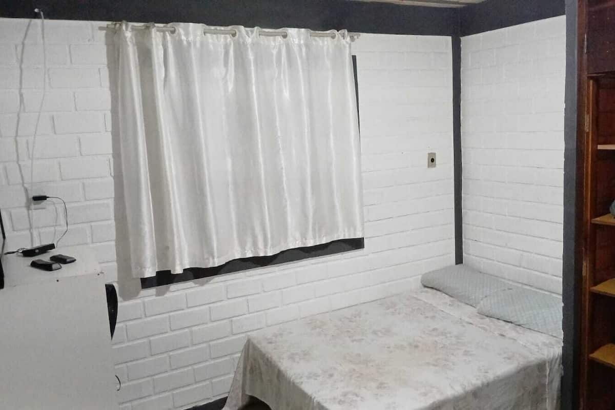 Ein kleiner, kompakter Raum mit weißer Backsteinwand, einem Fenster mit weißen Vorhängen, einem Bett mit weißer Bettdecke und einer Holzregaleinrichtung mit gefalteter Kleidung.