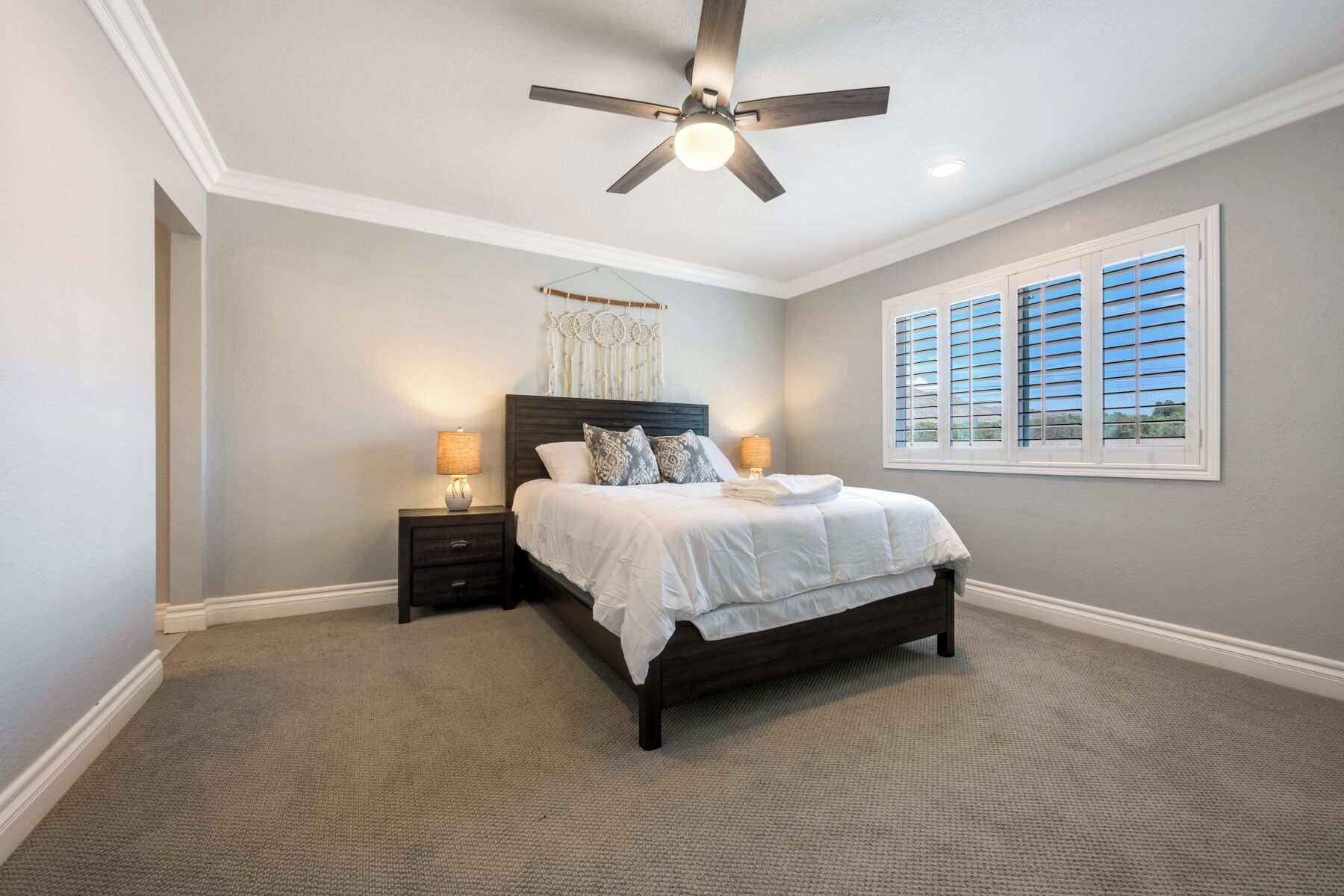 A bedroom with a dark wooden bed, two bedside lamps, a ceiling fan, and a window with blinds.
