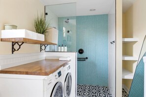 Un baño pequeño con lavadora, una repisa con una planta y una zona de ducha con azulejos estampados.
