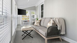 Un salon moderne avec un canapé, une petite table et un fauteuil en osier. Un téléviseur est fixé au mur, et il y a une fenêtre avec des stores.