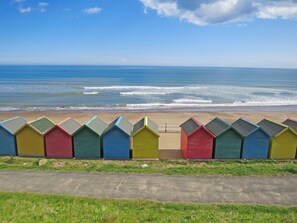 Une rangée de cabanes de plage colorées s’étend le long d’une plage de sable, avec l’océan en arrière-plan.