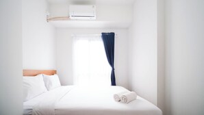 Ein Hotelzimmer mit Bett, weißen Laken, einem Fenster mit blauen Vorhängen und einer Klimaanlage.