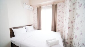 Quarto de hotel com cama, roupa de cama branca, cabeceira de madeira, cortinas florais e aparelho de ar condicionado.