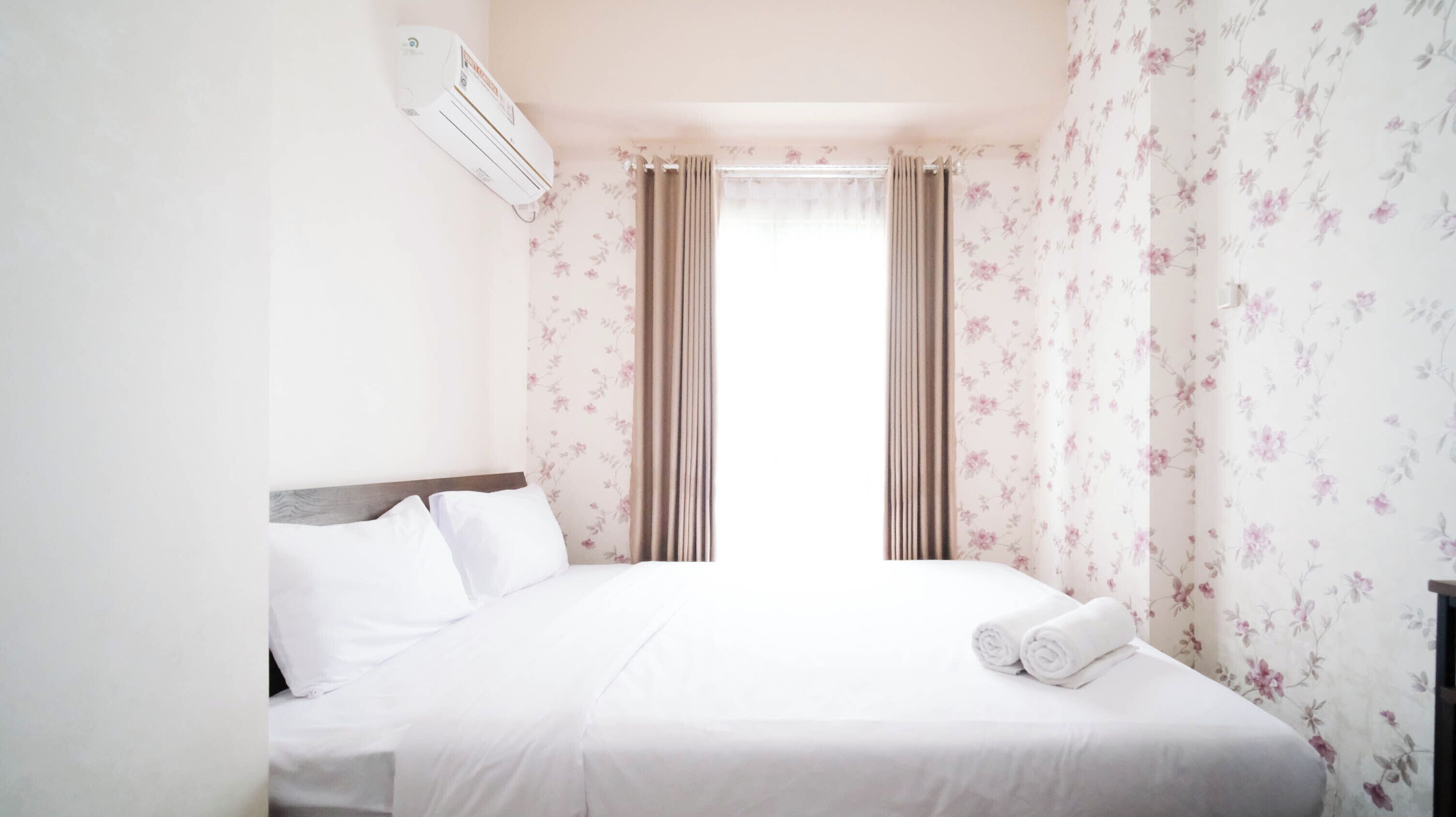 Een hotelkamer met een bed, witte beddengoed, bloemenbehang en een raam met gordijnen.