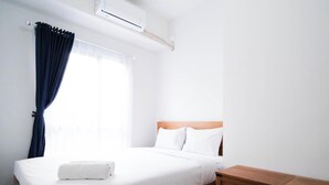 A neatly made bed with white linens and a rolled towel, a wooden nightstand, a window with a curtain, and a wall-mounted air conditioning unit. - Tidy And Cozy 1Br At Tamansari Prospero Sidoarjo Apartment (Sidoarjo)