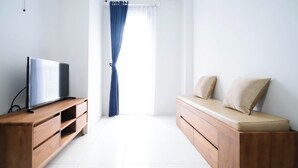A room with a wooden TV stand, a sofa with cushions, and a window with curtains. - Tidy And Cozy 1Br At Tamansari Prospero Sidoarjo Apartment (Sidoarjo)