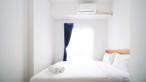 Una camera d'albergo con un letto, lenzuola bianche, un asciugamano arrotolato, una finestra con tende azzurre e un condizionatore d'aria.