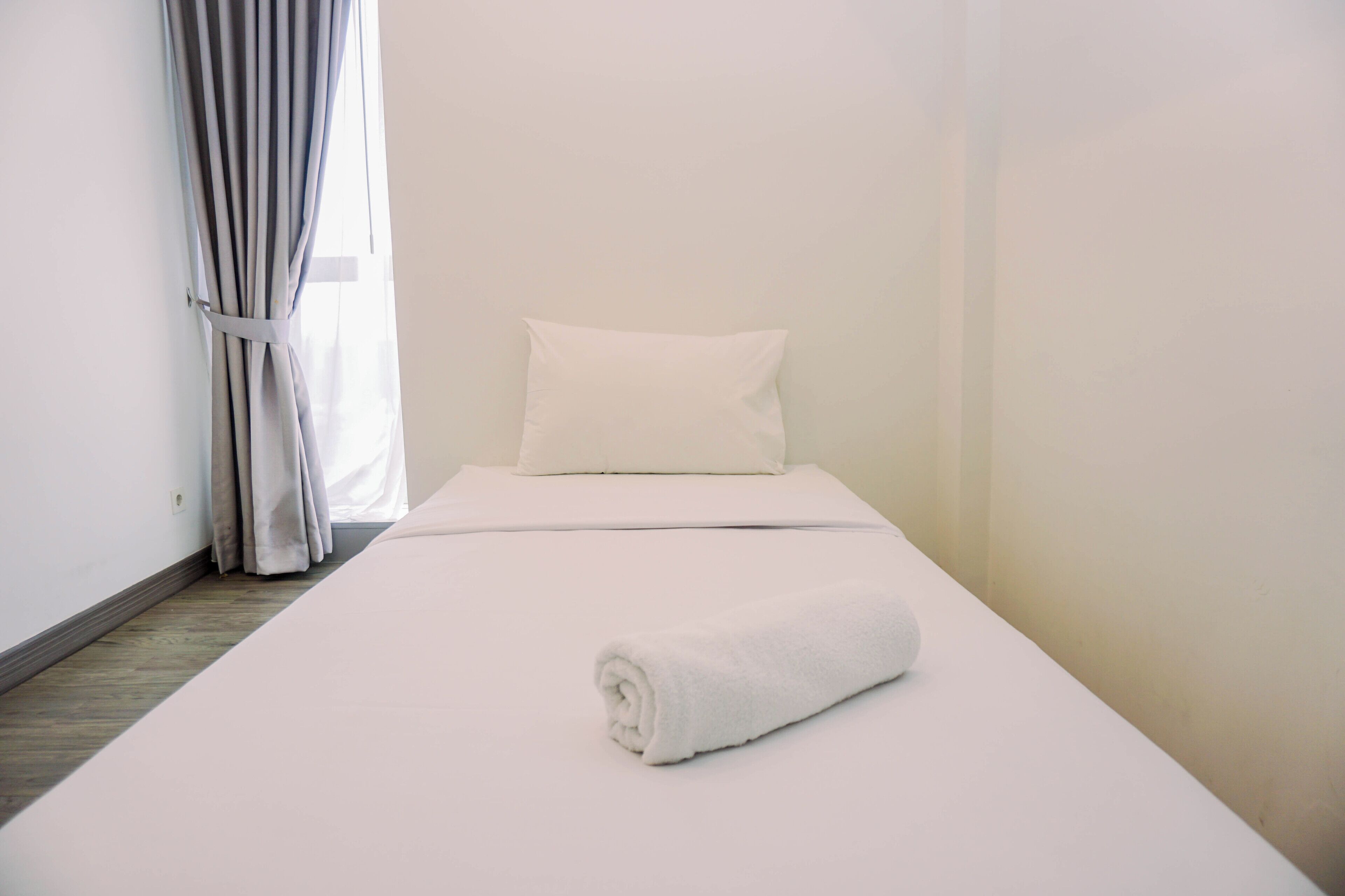 A single bed with a white pillow and a rolled white towel, a window with white curtains, and a wooden floor.