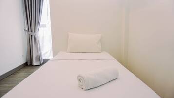 A single bed with a white pillow and a rolled white towel, a window with white curtains, and a wooden floor.