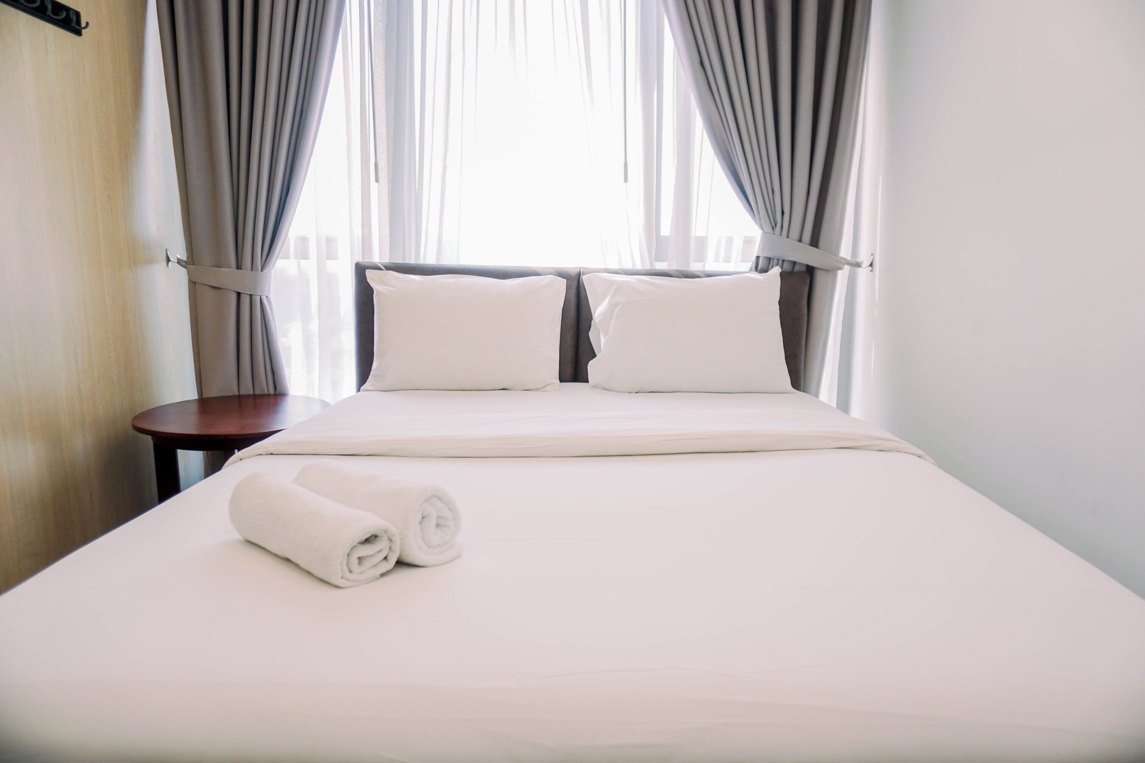 A neatly made bed with white linens and pillows, a small round table beside it, and sheer curtains on the window.