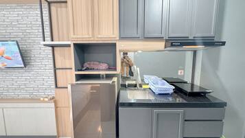 A modern kitchen with grey cabinets, a black countertop, and a built-in oven. There is a wooden shelf above the sink and a television mounted on the wall.