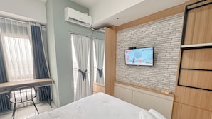 A hotel room with a bed, a desk with a chair, a television mounted on a brick-patterned wall, and an air conditioning unit on the wall. - Comfortable Living Studio At Tamansari Prospero Apartment (Sidoarjo)