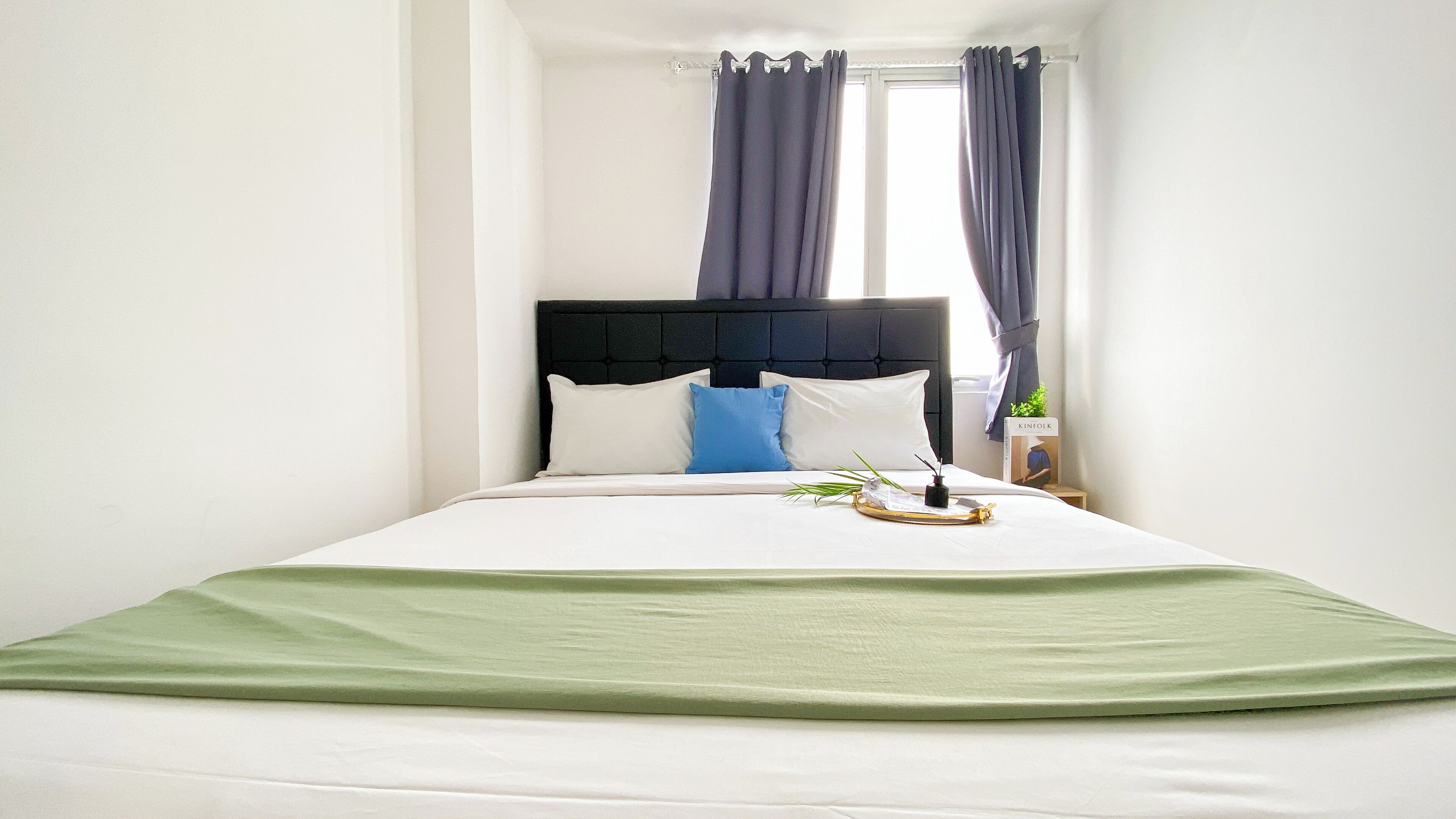 A neatly made bed with a green throw, white pillows, and a blue accent pillow. There is a small bedside table with a book titled "KINFOLK" and a plant.