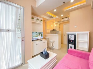 A modern hotel room with a TV, a minibar, a pink sofa, and a small kitchen area. - The Nifty Nook 2Br At Grand Asia Afrika Apartment (Bandung)