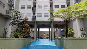 Una piscina con borde de azulejos, rodeada de vegetación y un edificio de varios pisos con balcones.