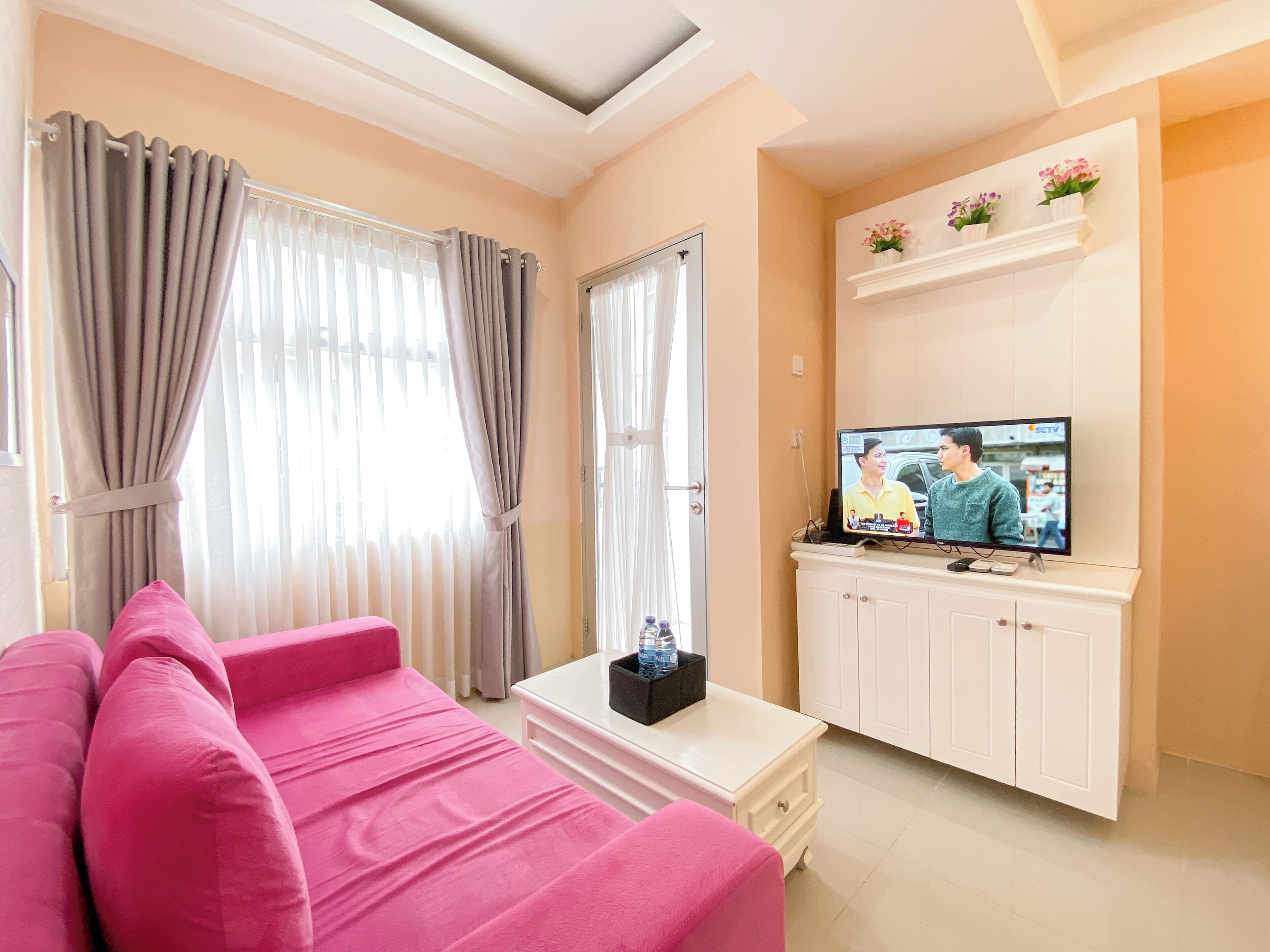Una sala de estar con un sofá rosa, un televisor sobre un mueble blanco y una ventana con cortinas.