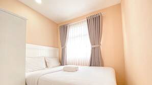 Una cama bien hecha con sábanas y almohadas blancas, un armario blanco y una ventana con cortinas.