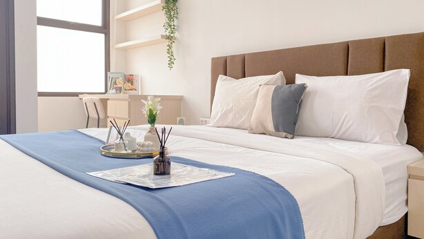 Ein ordentlich bezogenes Bett mit einer blauen Decke, weißen Kissen und einem Tablett mit einer Vase und Kerzen. Es gibt einen Schreibtisch mit einem Stuhl und eine Pflanze an der Wand.