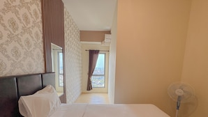 A hotel room with a large bed, a bedside table, a mirror, a window with curtains, and a standing fan. - Simply And Good Place 2Br At Tamansari Prospero Apartment (Sidoarjo)