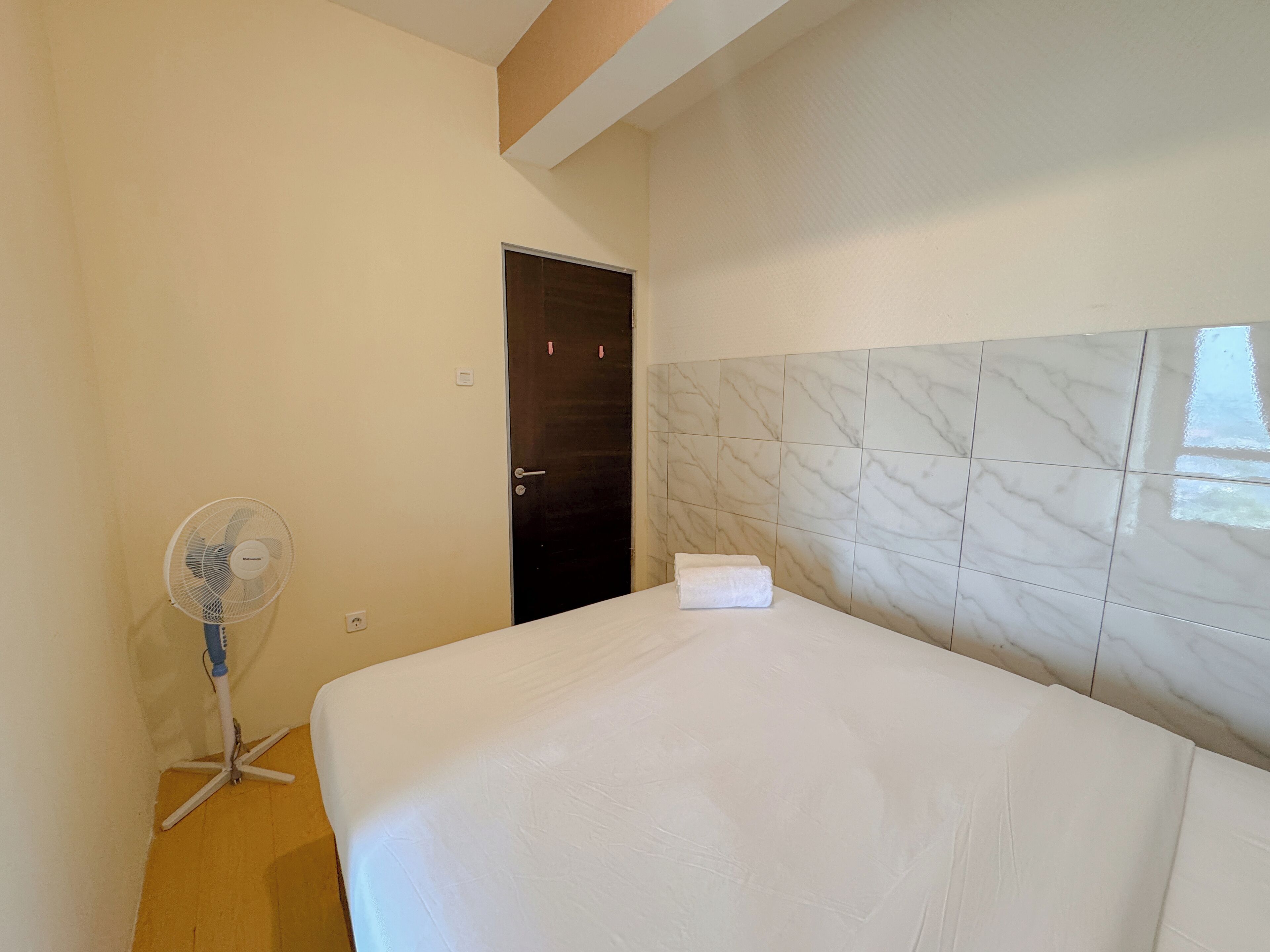 A hotel room with a bed, a standing fan, and a door.