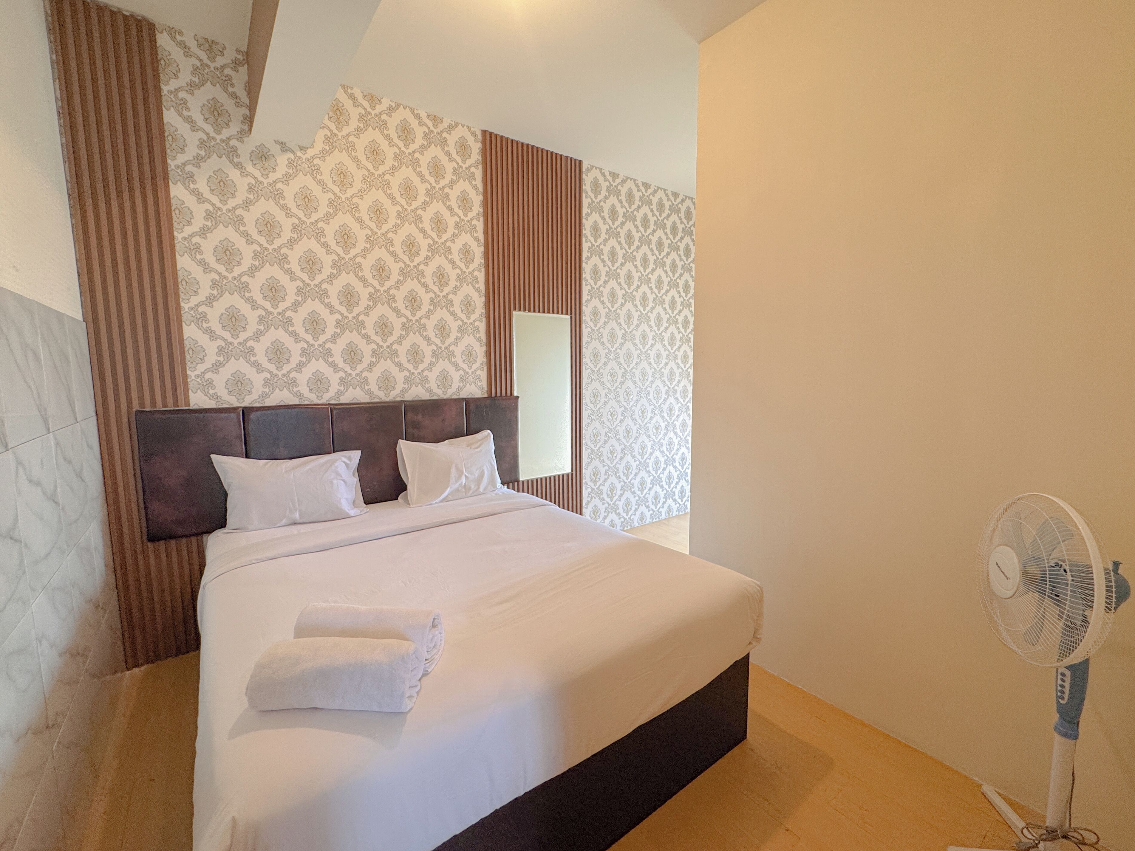 A neatly made bed with white linens and pillows, a bedside fan, and patterned wallpaper.